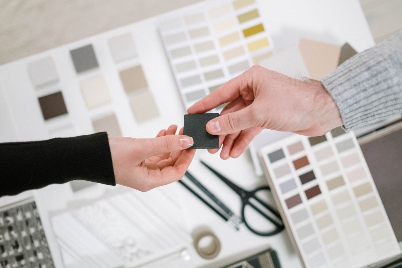 Two hands exchange a color sample during a collaborative design meeting with various palettes.